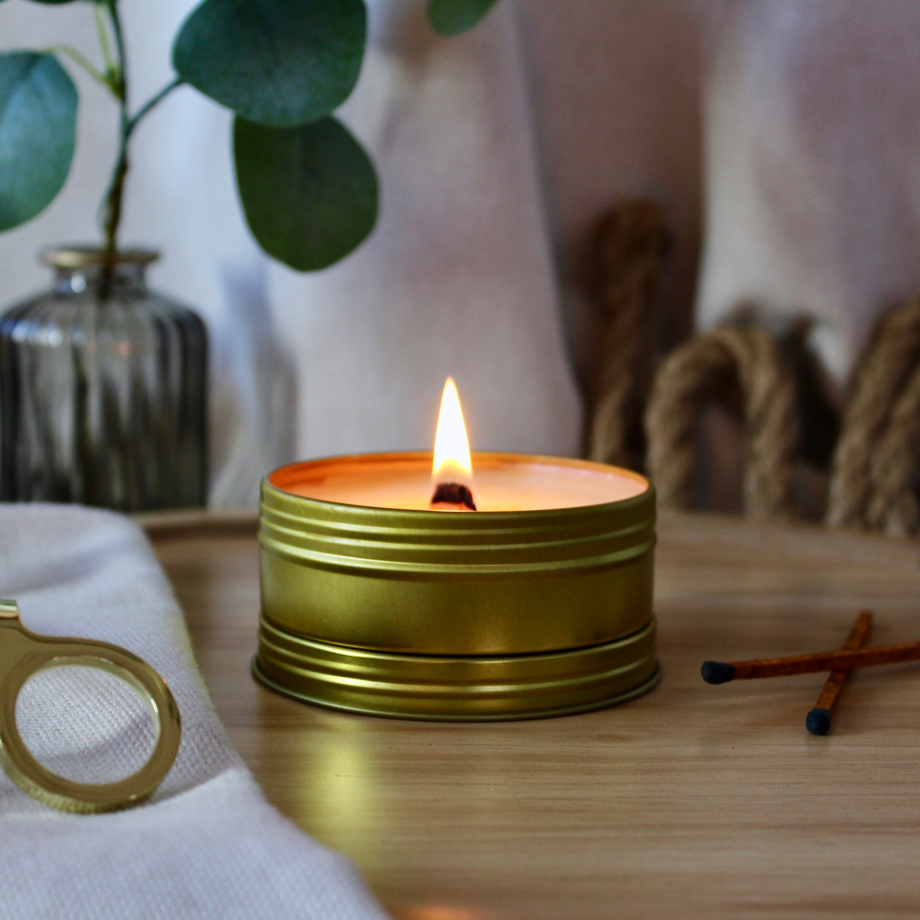 A lit travel candle in a gold tin container with a crackling wood wick. The container is placed on a wooden surface, and there is a blurred background featuring a plant and some decorative items.